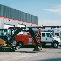 Pelleteuse compacte jaune et noire stationnée devant un bâtiment industriel avec plusieurs camions utilitaires alignés à côté.