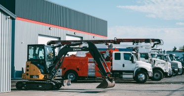 Pelleteuse compacte jaune et noire stationnée devant un bâtiment industriel avec plusieurs camions utilitaires alignés à côté.