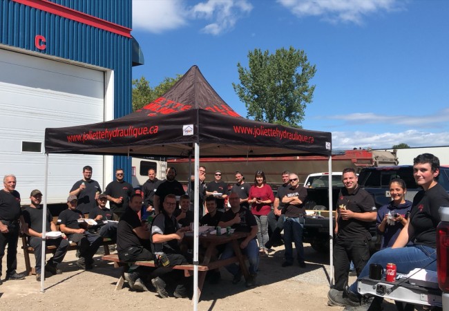 Groupe d'employés en tenue noire partageant un repas sous un chapiteau noir devant un bâtiment industriel par temps ensoleillé.