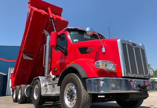Camion benne dompeuse rouge avec benne levée, stationné devant un bâtiment bleu sous un ciel dégagé.