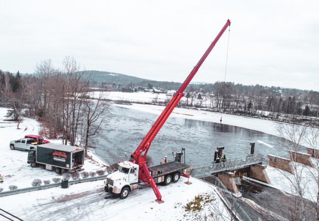 Camion-grue rouge avec système hydraulique en opération près d'un pont sur une rivière en hiver enneigé.
