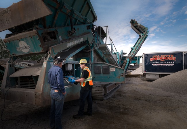 Deux travailleurs discutent près d'une machine industrielle bleue sur un chantier avec un camion de Joliette Hydraulique en arrière-plan.