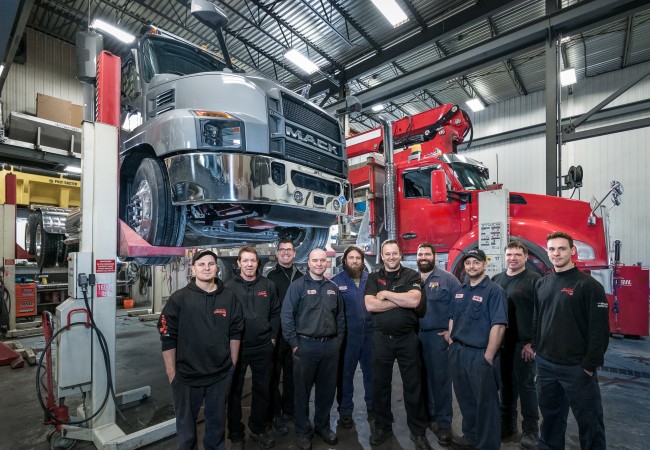 Équipe de mécaniciens posant dans un atelier avec un camion Mack gris sur un pont élévateur et un camion rouge en arrière-plan.