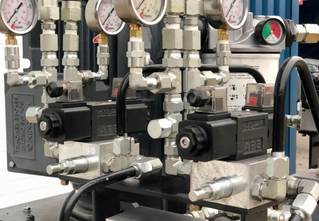 Pompe hydraulique avec plusieurs manomètres et vannes connectées par des tuyaux noirs sur un support métallique.