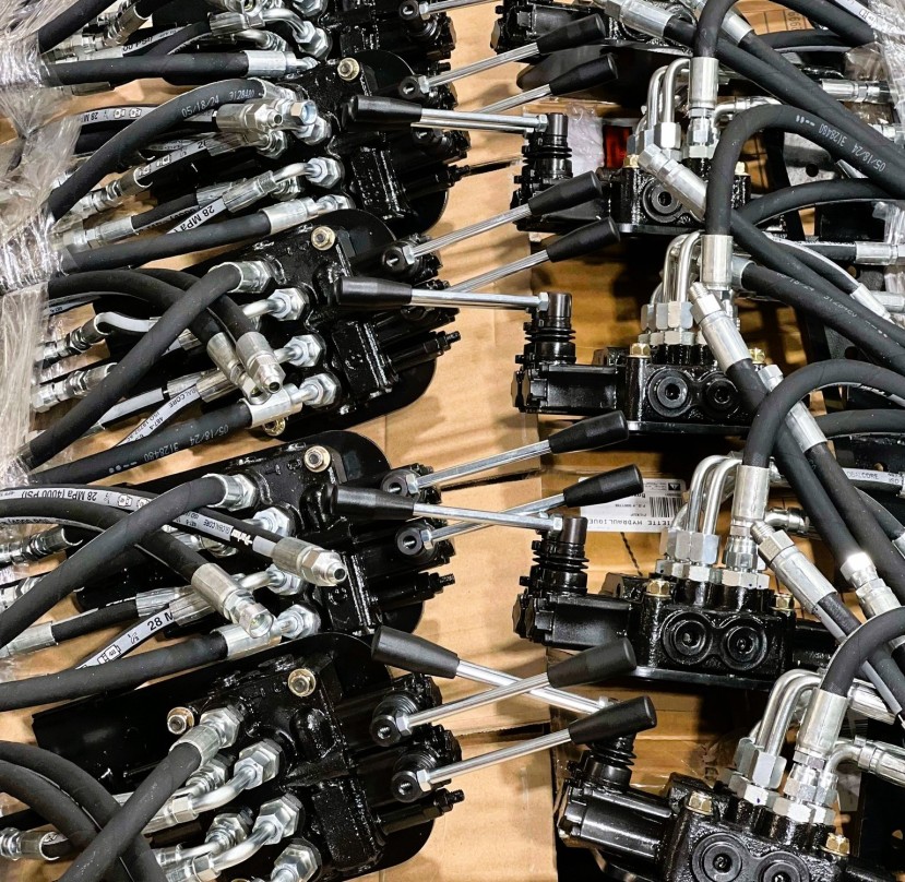 Plusieurs valves hydrauliques noires avec leviers métalliques et tuyaux flexibles sont disposées sur une surface en carton.